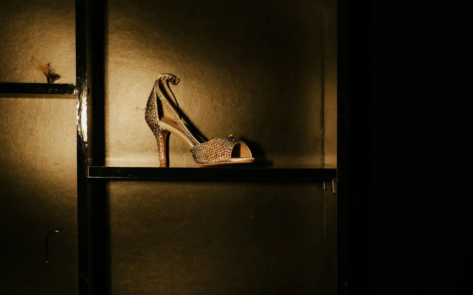 Glitzernde Riemchensandalette mit Absatz auf einem dunklen Regalbrett, seitlich beleuchtet